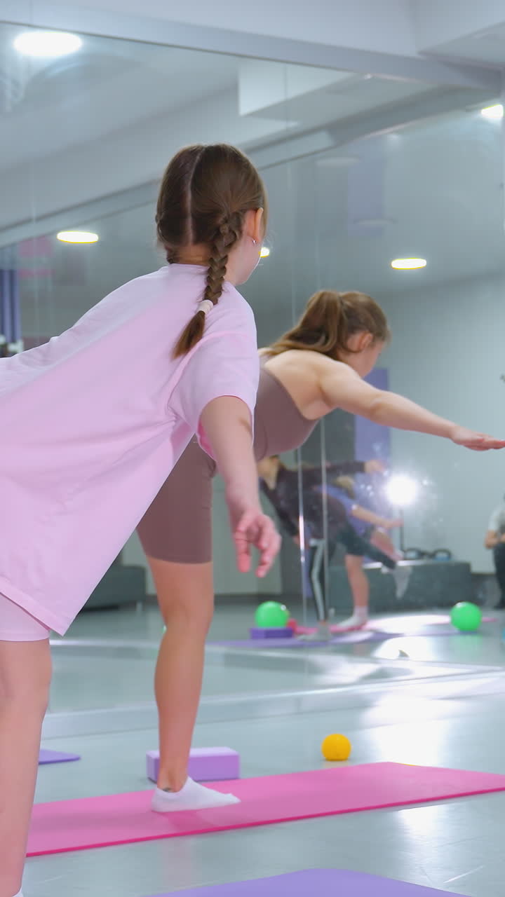 Sessione di allenamento di gruppo in una luminosa sala fitness con partecipanti che si bilanciano su una gamba sola con le braccia aperte, palle da ginnastica colorate sparse intorno, un grande specchio che riflette i loro movimenti.