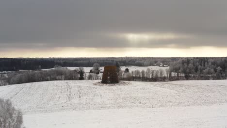 Aerial drone shot of an old windmill in the middle of a field covered with snow. Winter day with sun rays breaching through the clouds.
