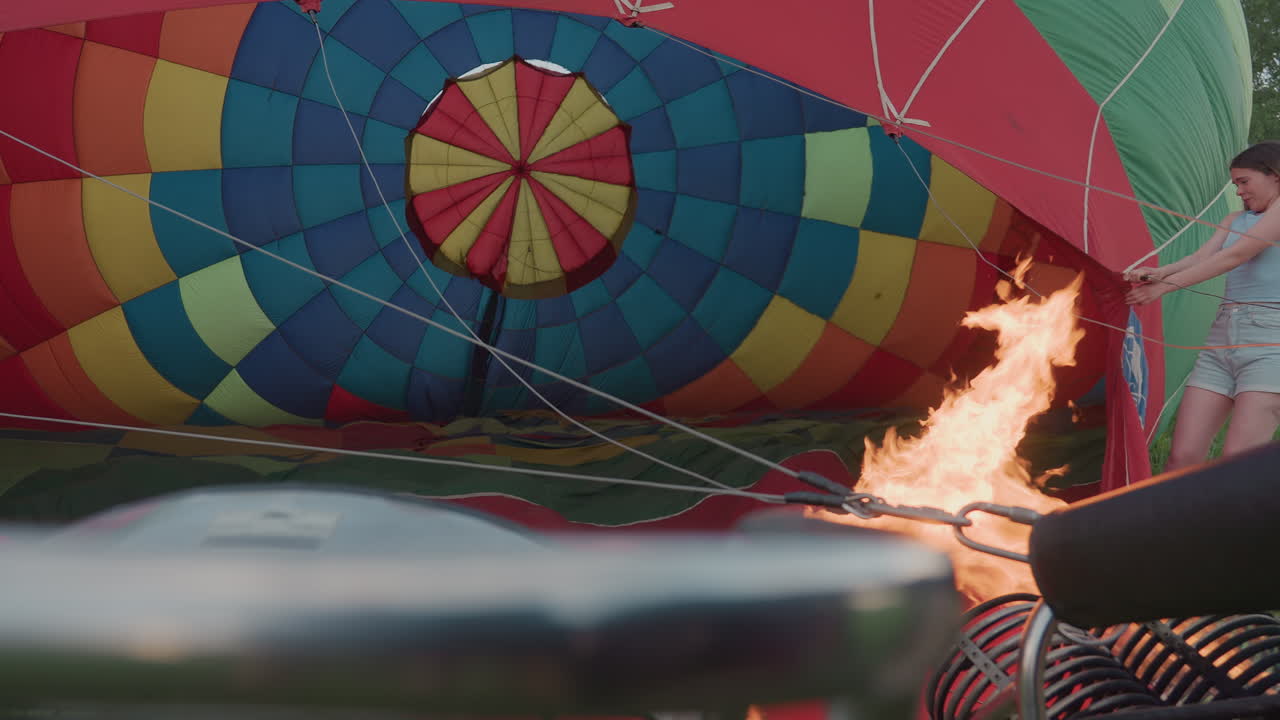 close up of metal burner flame shooting into colorful hot air balloon envelope while operator pulls fabric open in tall grass field during sunset preflight inflation detailed lift off prep