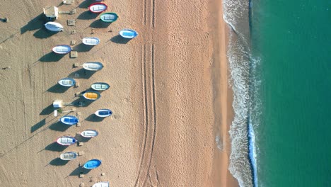 overhead view showing vibrant boats neatly aligned on a serene sandy beach next to turquoise sea waves gently reaching the shore