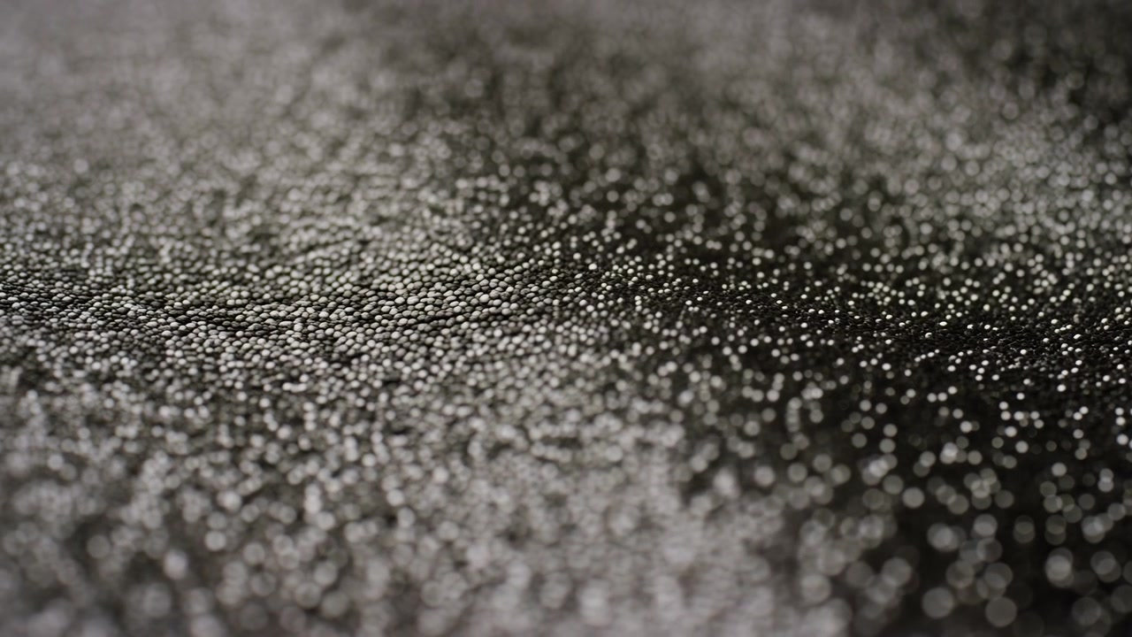 Inúmeras pequenas gotas de água cobrem um fundo texturizado escuro em profundidade de campo rasa, criando um padrão abstrato cintilante com realces suaves.