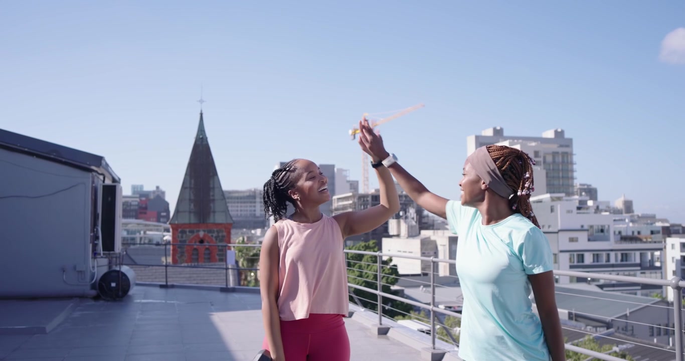 Two African women starting set with hex dumbbells overhead, doing synced squats on rooftop training