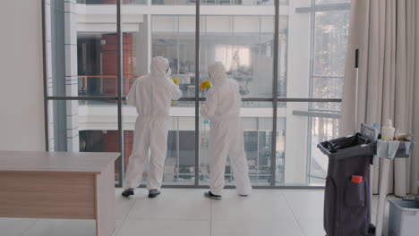 Rear-View-Of-Two-Cleaning-Men-Wearing-Personal-Protective-Equipment-Cleaning-Crystal-Walls-Inside-An-Office-Building