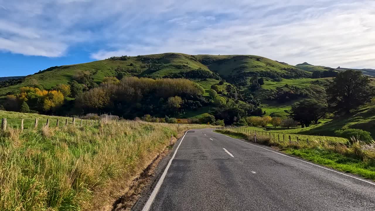 A forward-moving camera travels along a rural road bordered by grassy fields and rolling green hills under bright daylight and partly cloudy skies