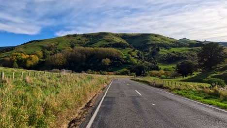 a forward-moving camera travels along a rural road bordered by grassy fields and rolling green hills under bright daylight and partly cloudy skies