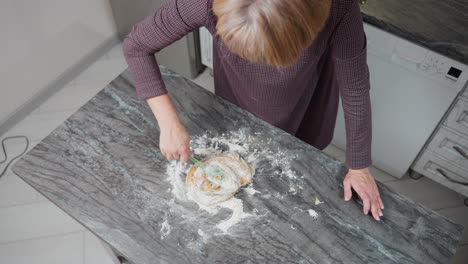 overhead view of baker kneading soft dough on floured tiled table while resting left hand, kitchen workspace covered in flour as homemade baking process unfolds