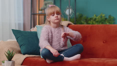 Child-girl-kid-sitting-on-sofa-at-home-alone-showing-thumbs-down-sign-gesture,-dislike,-disapproval