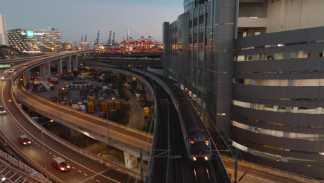 mtr commuter train overpassing highway with industrial port in the background after sunset