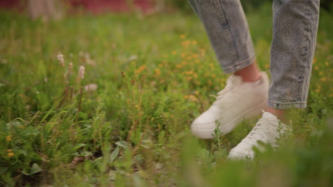 close-up of young woman s legs in light denim jeans and white sneakers walking through grassy field, with lush green grass and gentle movement as she steps forward