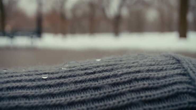 Clear beads remain on raised rows of gray yarn while closer framing emphasizes rounded reflections and soft fibers, with snow, bare tree trunks and distant bench blurred behind in muted daylight.