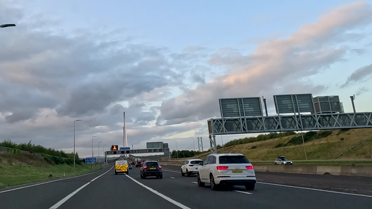 Vehicles travel under highway signs during a picturesque evening drive with a view of the sky and clouds.