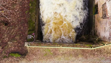 Powerful-man-made-waterfall-near-abandoned-millhouse-building,-aerial-top-down-view