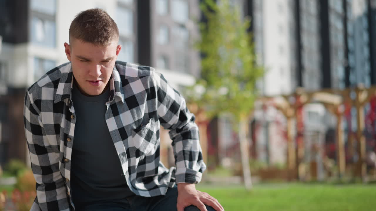 young sick student in checkered shirt leans forward while retrieving medicine from pocket in outdoor park setting with tall residential buildings and blooming flowers in soft background