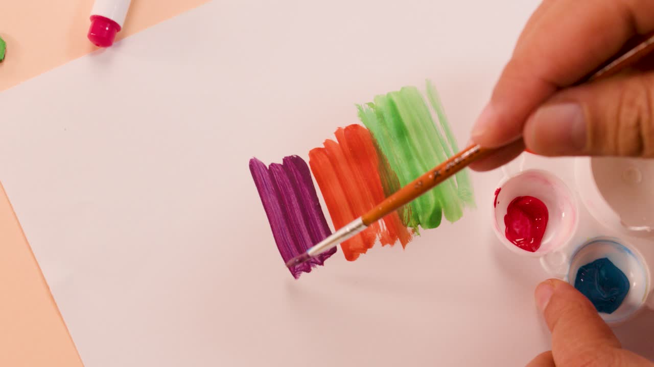 Hand paints vibrant acrylic stripes on paper under soft lighting, overhead view, pastel background