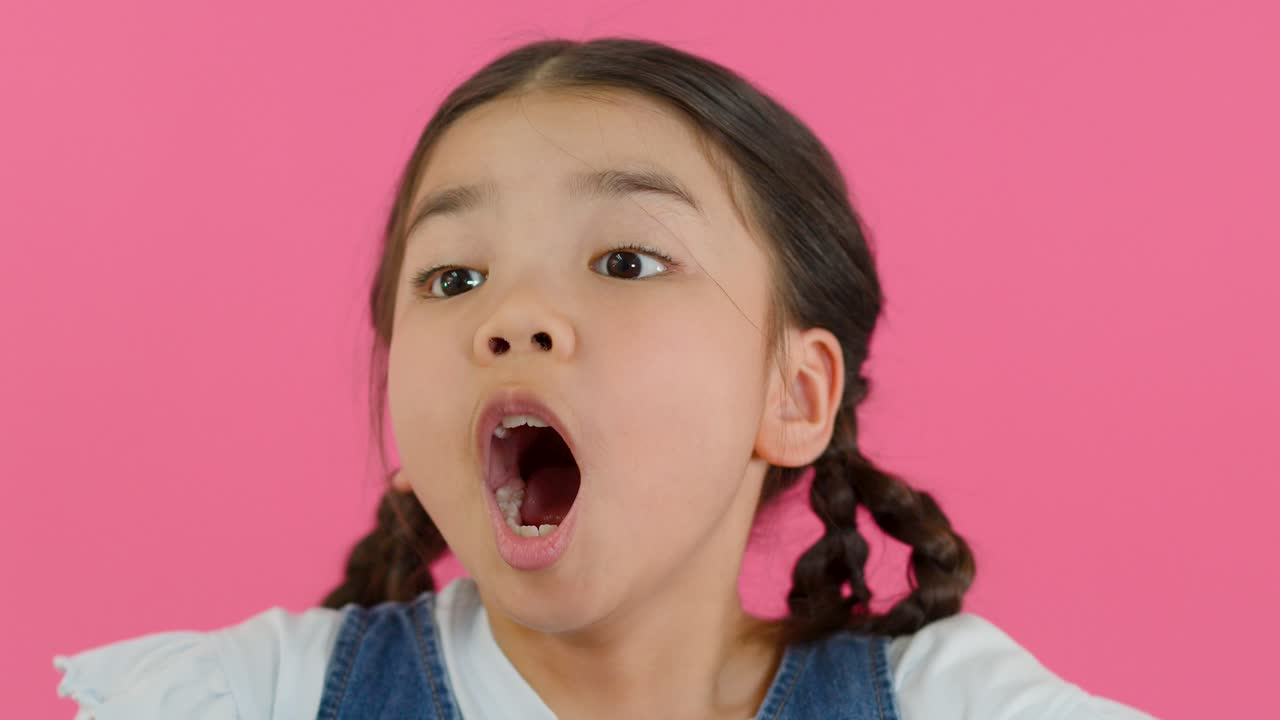 Bambina con codini reagisce con sorpresa, mani vicino al viso, illuminazione da studio brillante, sfondo rosa