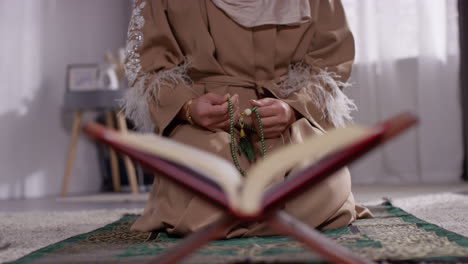 Close-Up-Of-Open-Copy-Of-The-Quran-On-Stand-With-Muslim-Woman-Praying-With-Prayer-Beads-In-Background