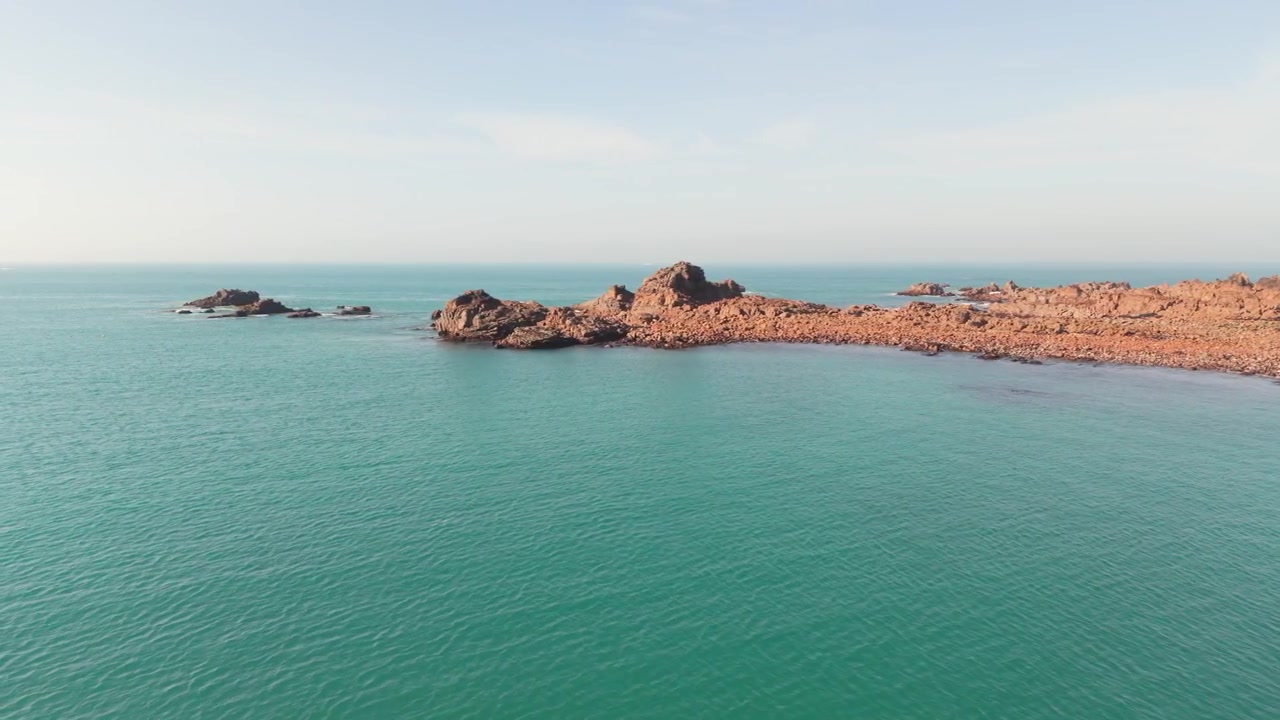 Droneoptagelse af en klippefyldt kystlinje, der langsomt bevæger sig mod havet, med et roligt, klart hav i den sene eftermiddagssol, mod granitfremspring, under en klar blå himmel og med udsigt til horisonten