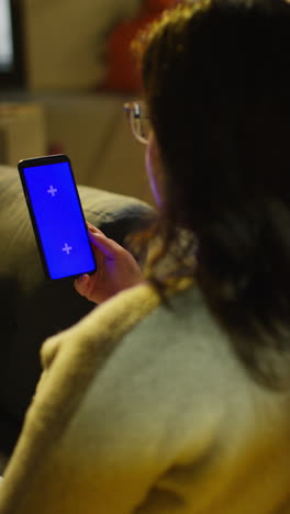 Vertical-Video-Close-Up-Over-The-Shoulder-Shot-Of-Woman-Spending-Evening-At-Home-Sitting-On-Sofa-Looking-At-Blue-Screen-Mobile-Phone