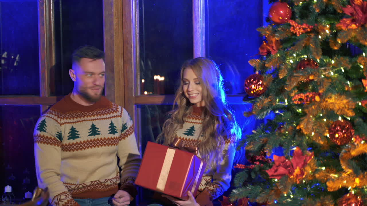Man is giving a woman Christmas present. Happy beautiful girl takes a box with present. Couple in love near the Christmas tree. New year atmosphere at home.