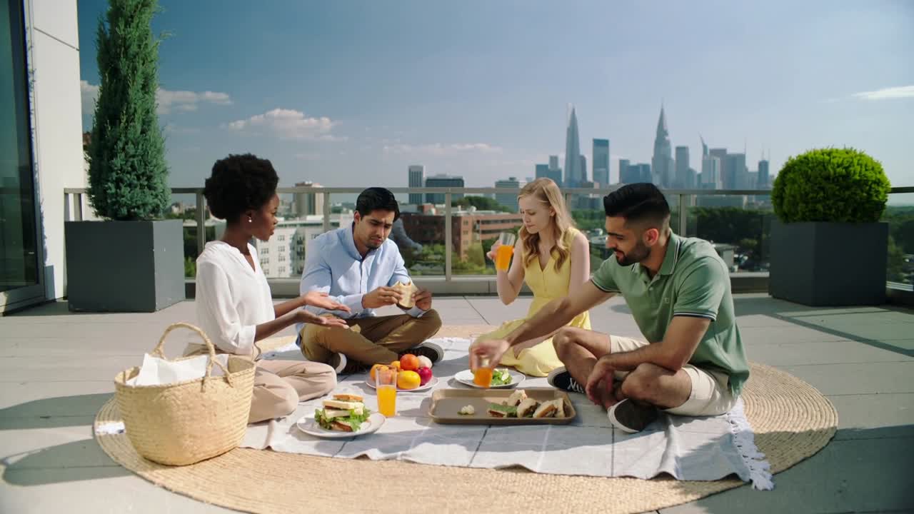 Offering woman in white blouse passing sandwich on rooftop rug, friends sipping juice for picnic