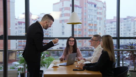 team leader standing in front of the table and speaking to his colleagues. office workers at the table discussing ideas. city scene at the background.