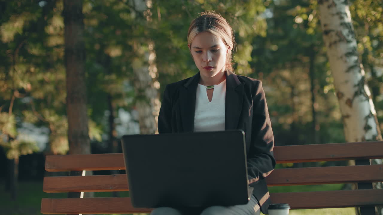 Focused remote worker picks up drink and sips from it while working on laptop outdoors with sunlight highlighting her face surrounded by trees and greenery