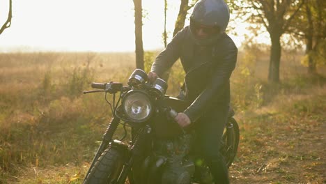 young man in helmet and leather jacket comes up to his bike and starts the engine while standing on the roadside in a sunny day