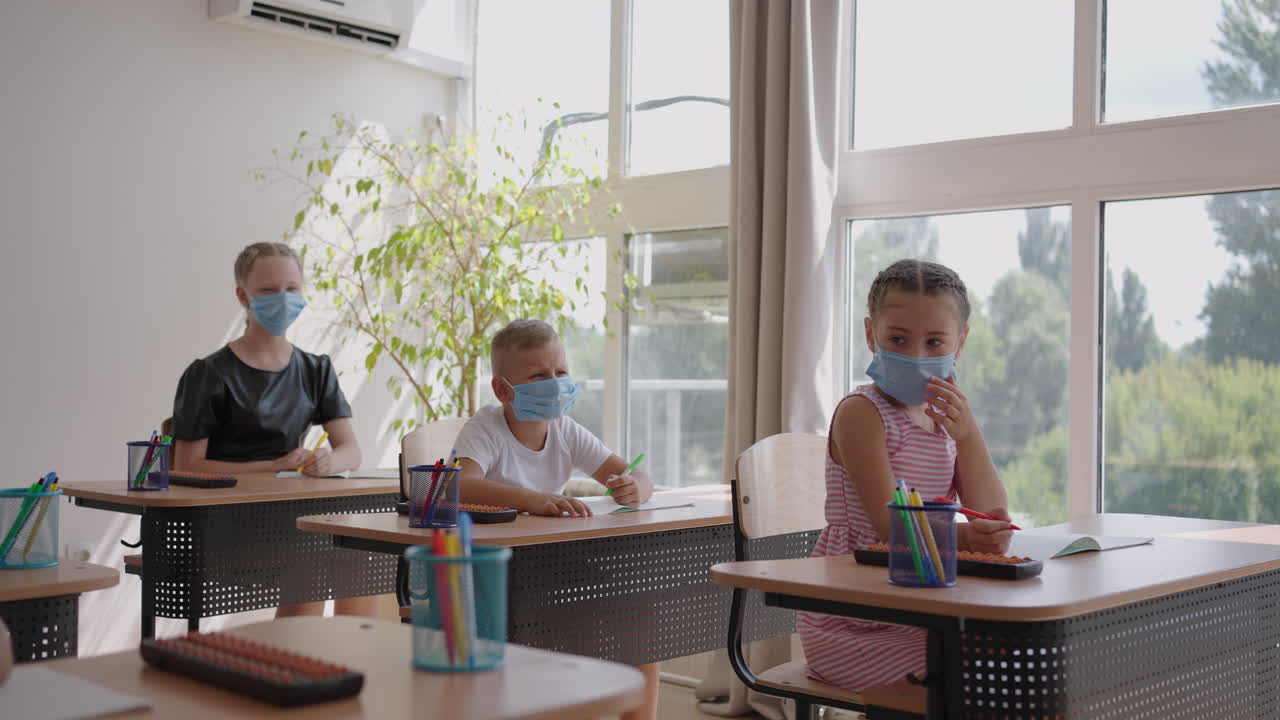 학교에서 아이들은 교실에 앉아서 손을 들고 스로모션으로 보호 마스크를 착용하여 교사의 질문에 대답합니다.