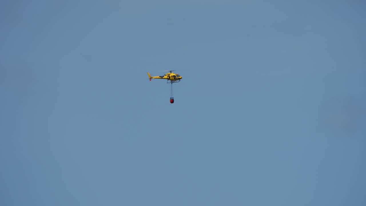 Yellow helicopter flying in clear blue sky carrying red bambi bucket for firefighting