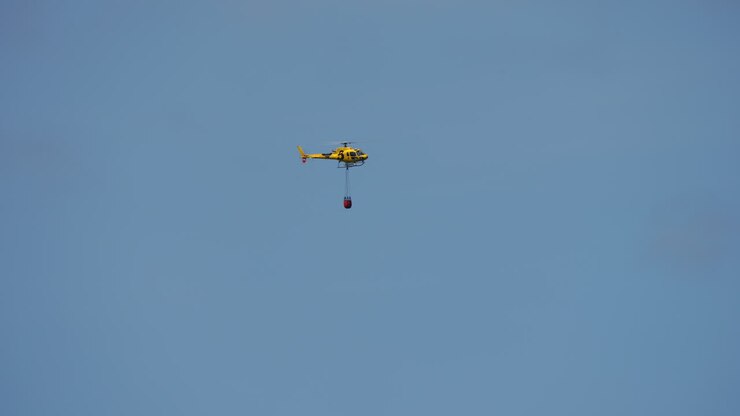 Yellow helicopter flying in clear blue sky carrying red bambi bucket for firefighting