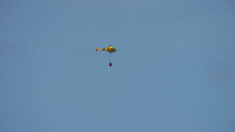yellow helicopter flying in clear blue sky carrying red bambi bucket for firefighting