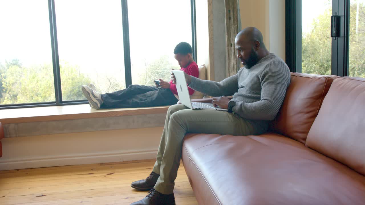 Afroamerikansk far og sønn som slår seg ned og deretter skriver på arbeids-laptop, scroller på telefonen i stuen