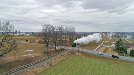 Una-Vista-Aérea-De-Un-Tren-De-Vapor-Que-Se-Acerca-Viajando-A-Través-Del-Campo-Soplando-Humo-Y-Vapor-En-Un-Día-De-Invierno