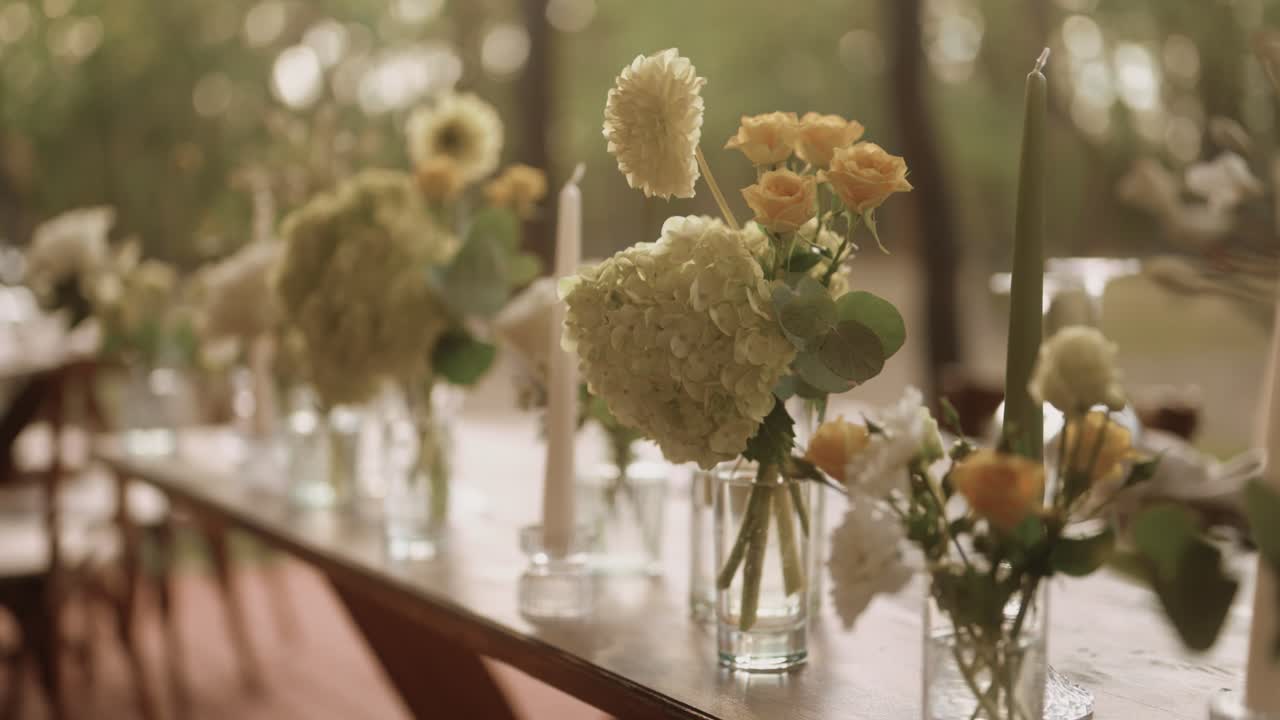 Close up shot of an elegantly decorated table captured, showing delicate floral arrangements in glass vases, tall candles, in warm natural light