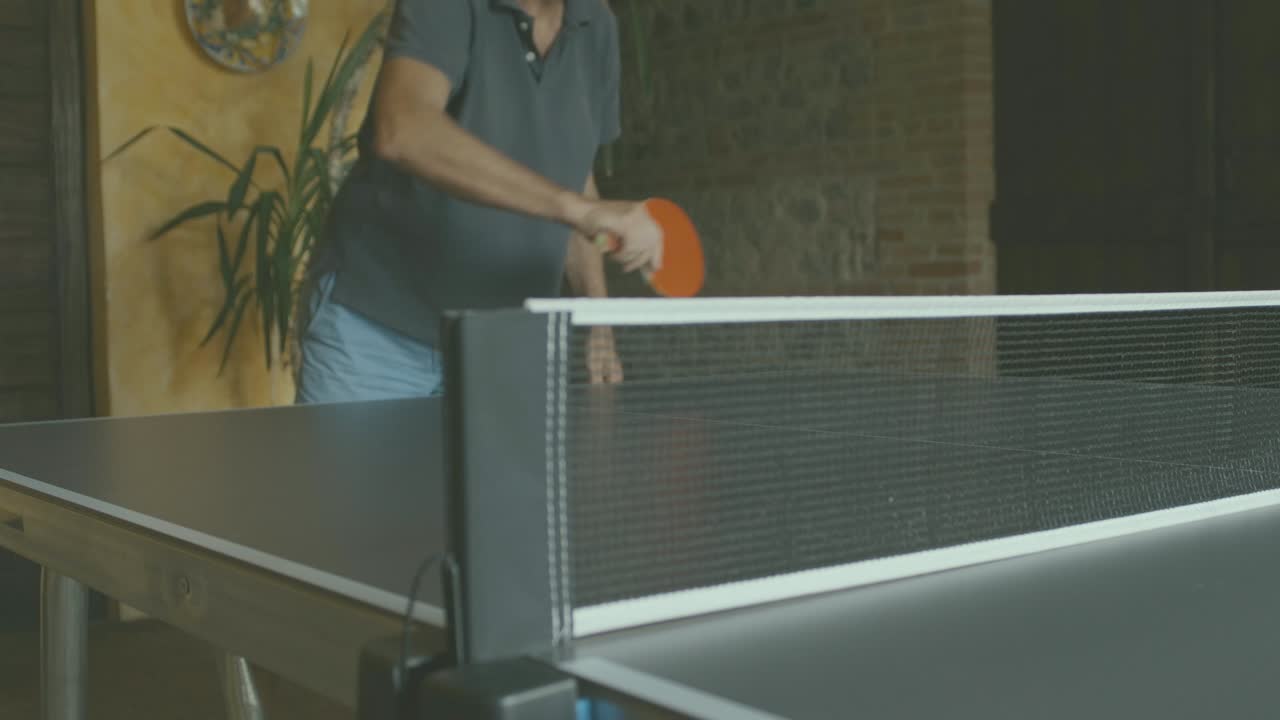 Un hombre está jugando al ping-pong, tomado desde abajo; se ve la red y, detrás de ella, una persona golpeando la pelota con una pala roja.