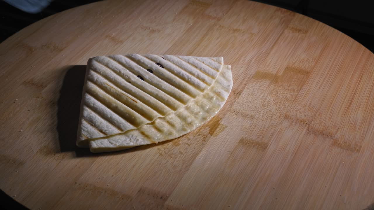 un primer plano de una quesadilla recién asada que se coloca sobre una tabla de cortar, el aceite del queso derretido gotea de la envoltura de tortilla tostada sobre la superficie de madera