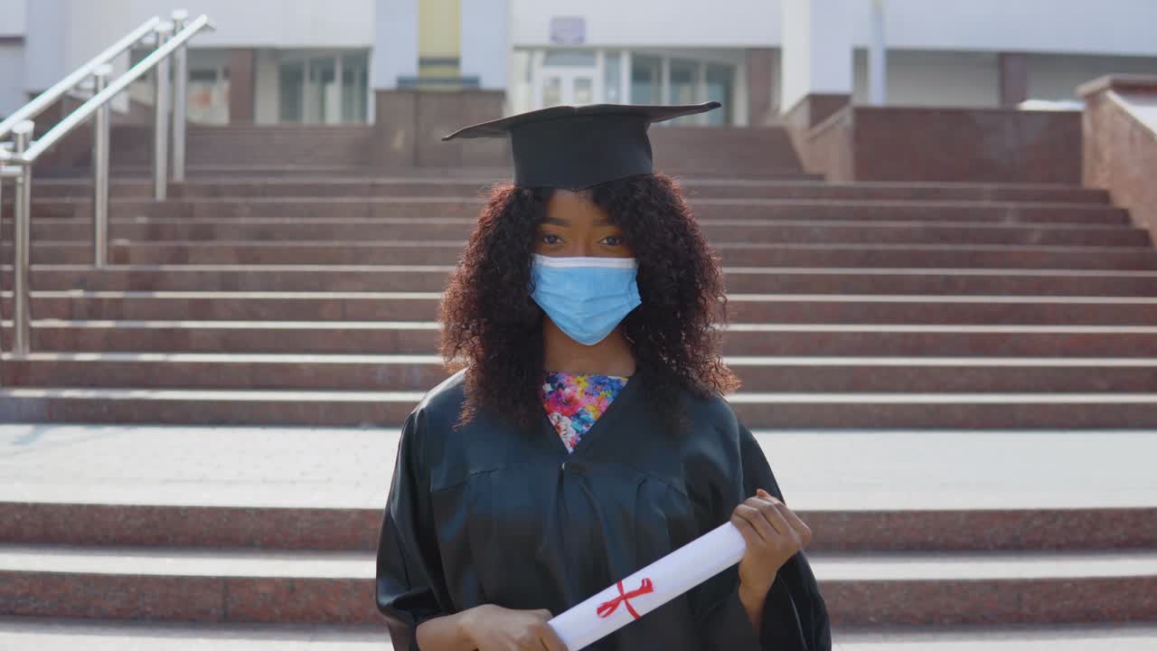 una joven afroamericana graduada con máscara médica protectora de pie frente a la cámara con un diploma en las manos. la estudiante está de pie en las escaleras afuera.