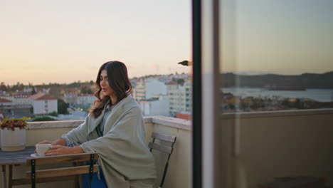 reflective woman sipping coffee on sunrise balcony illuminating cityscape alone