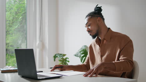 Busy-guy-writing-paper-data-in-light-office.-Afro-man-studying-looking-laptop