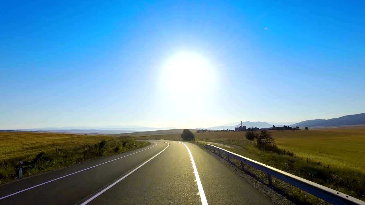 Drive on rural asphalt curved road among harvesting agriculture fields backlit by summer sun on blue sky, cinematic shot