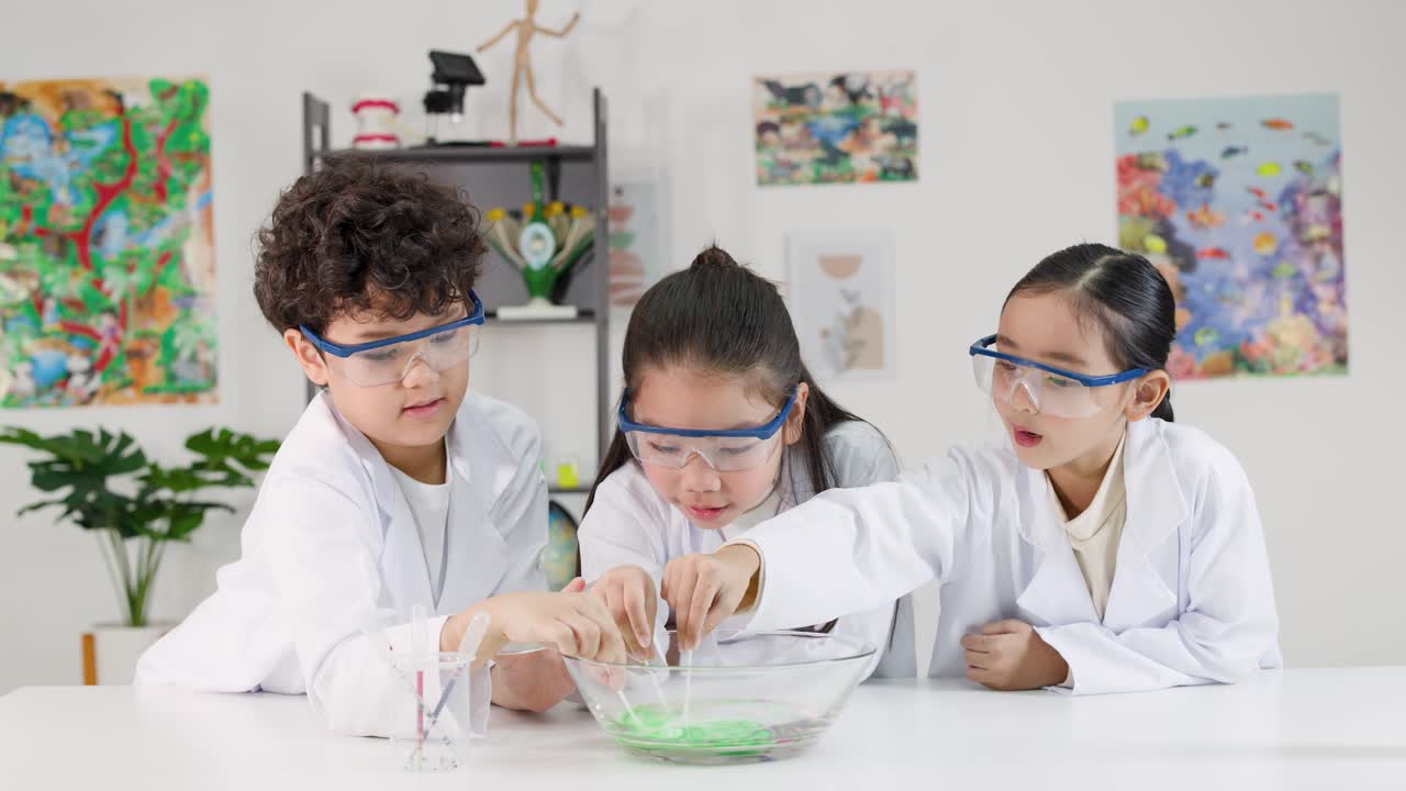 Tiga anak Asia dengan jas lab dan kacamata pelindung melakukan eksperimen susu berwarna-warni di kelas.