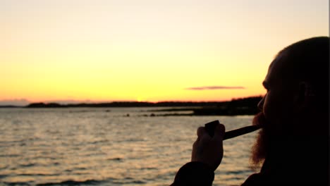 primer plano de un hombre con barba fumando una gruesa pipa liberando humo de su boca es movido detrás de él por el viento creado por las olas del agua el océano relajándose por sí mismo durante la hora del atardecer