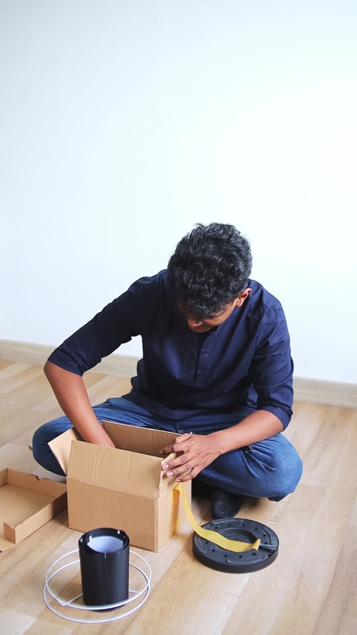 Young indian man unboxing lamp parts from open cardboard box on floor
