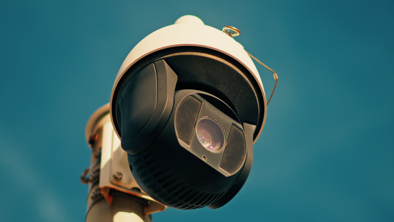 Close up of a round, black and white surveillance camera mounted on a white pole with the sky on the background