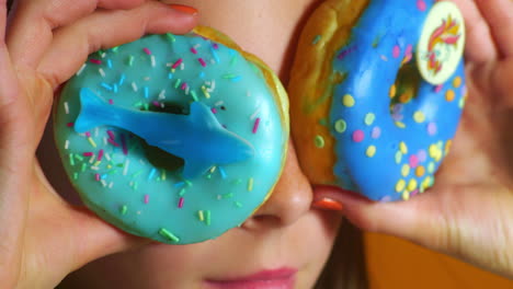 a child playfully holds two decorated blue donuts—one with a gummy shark, the other with a unicorn print and confetti sprinkles—over their eyes like glasses, creating a fun and imaginative scene.