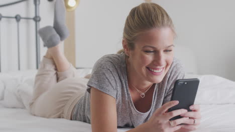 mujer joven sonriente acostada en la cama con teléfono móvil con pijama