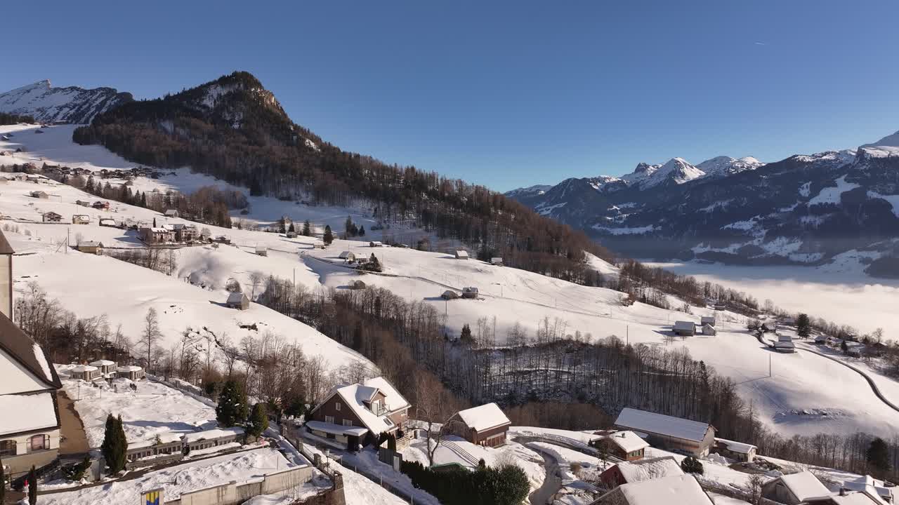 Aerial panorama above Amden, Switzerland, nestled amidst a pristine winter landscape. Snow-draped hills and rugged mountains frame this serene and majestic scene.
