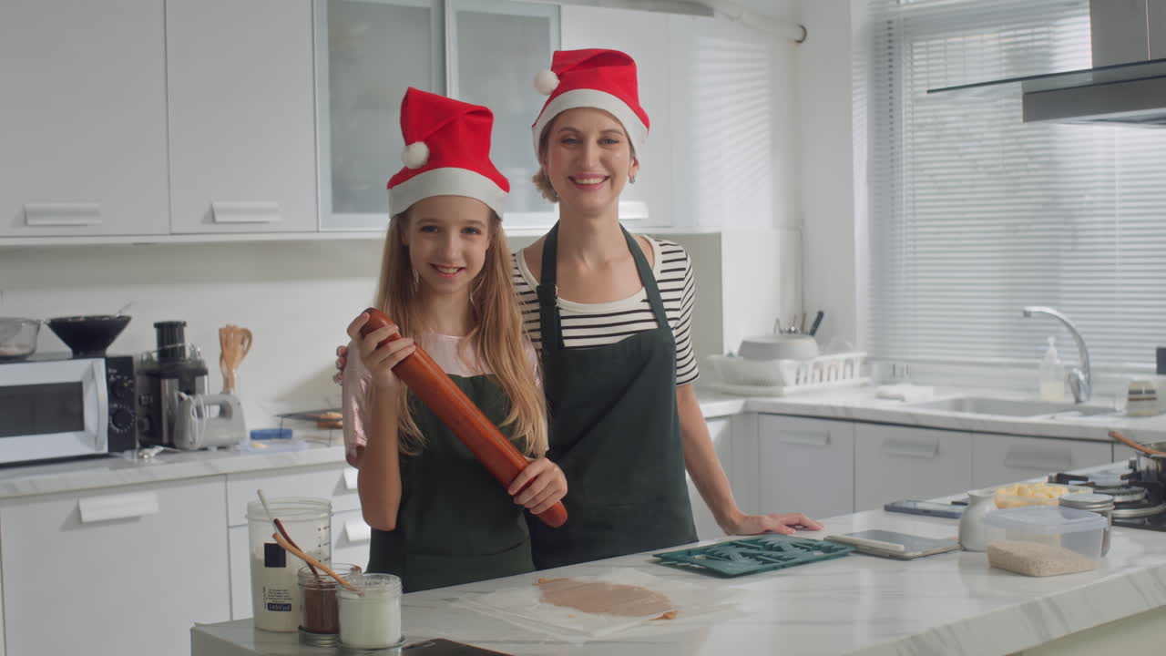 Positive Mom and Daughter in Christmas Hats Posing for Camera at Kitchen
