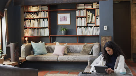 biracial woman sitting on sofa and using tablet at home, slow motion with copy space