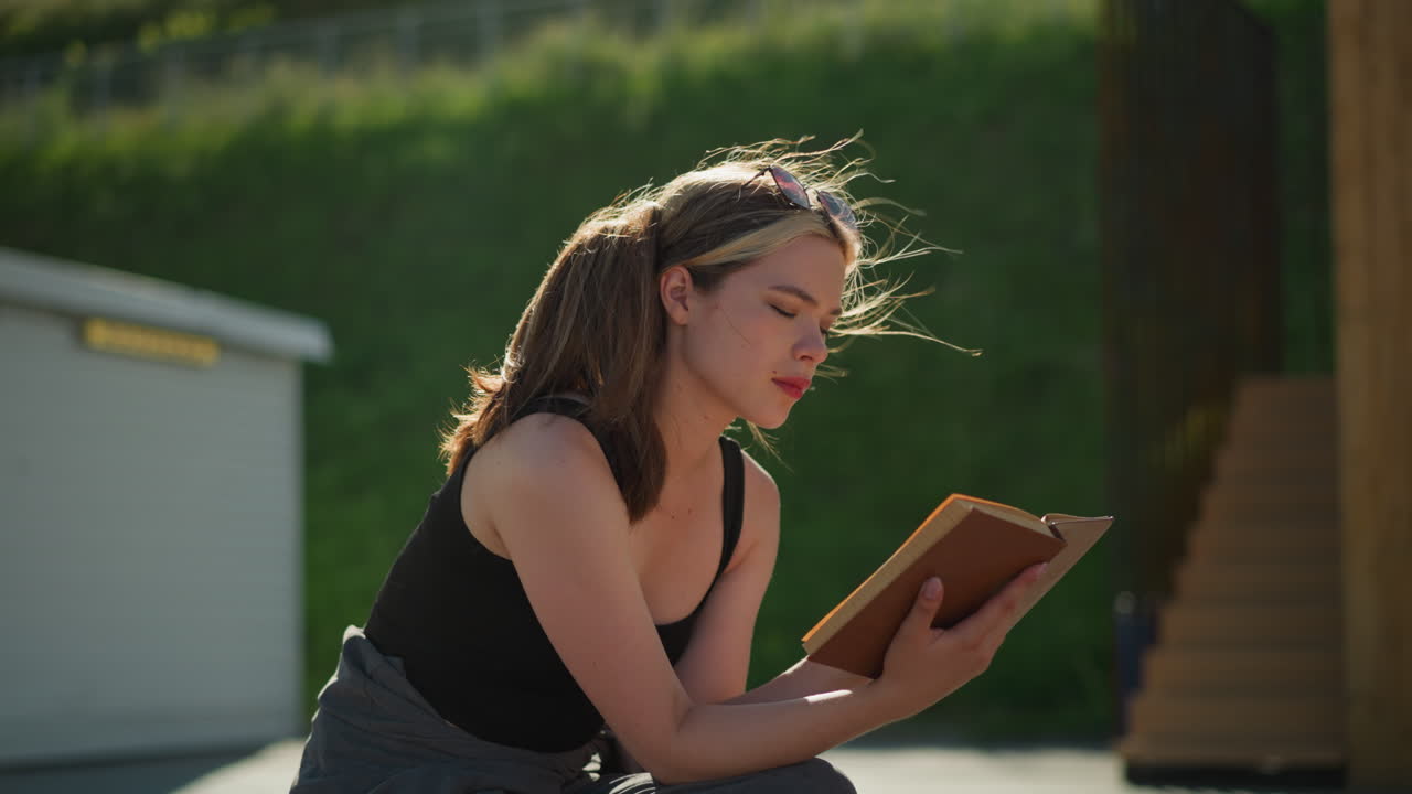 Lady seated outdoors reads a book while the wind blows her hair and lifts the book's pages, with sunlight reflecting on her, the blurred background features a staircase and greenery